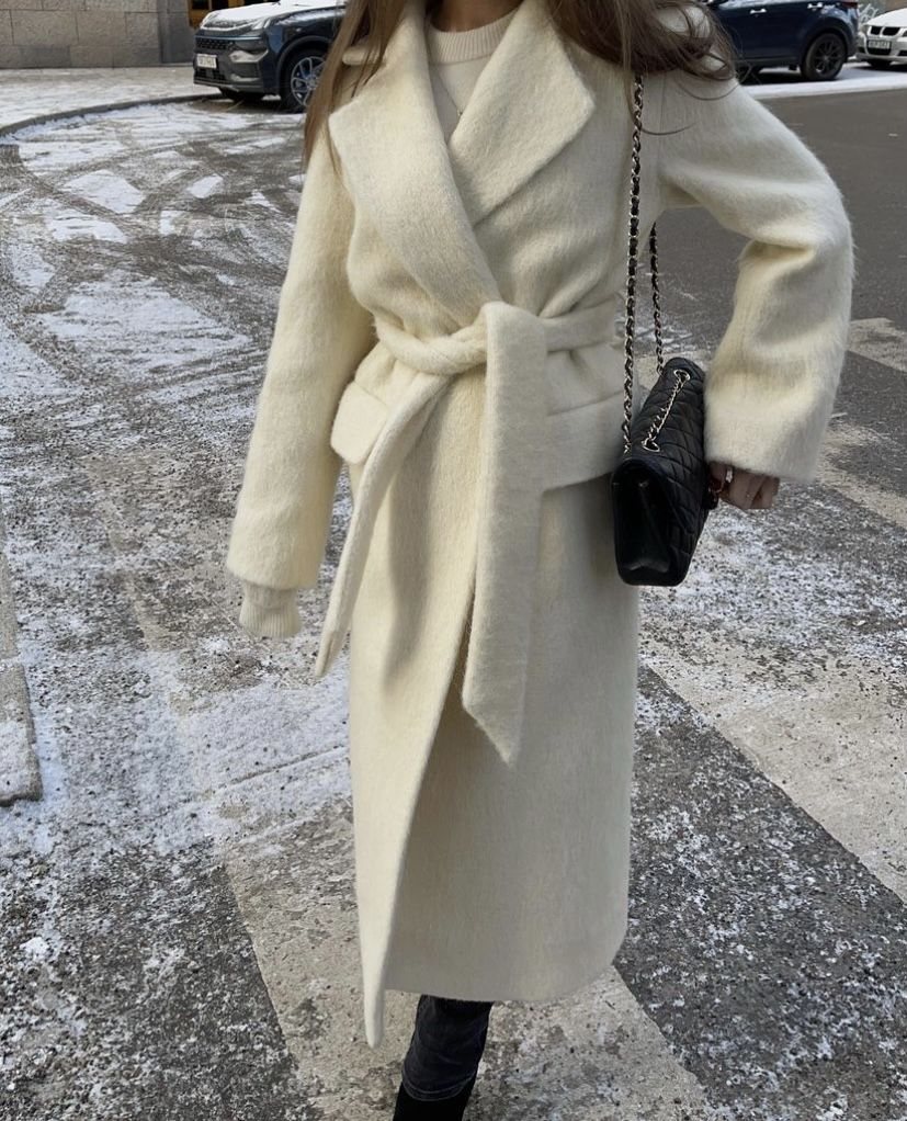 A woman walking in a snowy street wearing a long off-white belted wool coat.