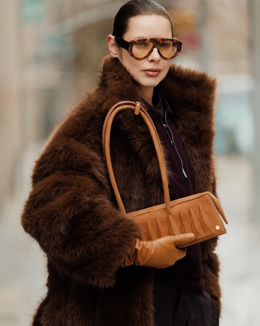 Woman in a brown fur coat wearing sunglasses, leather gloves, and carrying a tan handbag.