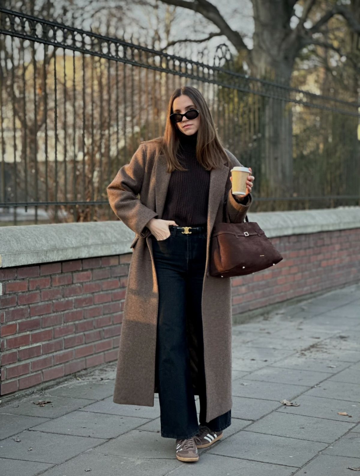 A woman in an oversized brown coat, dark turtleneck, and black trousers standing outdoors.