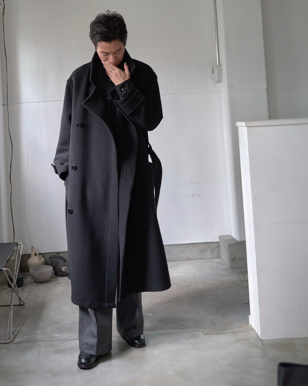 Man wearing an oversized black coat, black turtleneck, and grey tailored trousers standing in a white room.