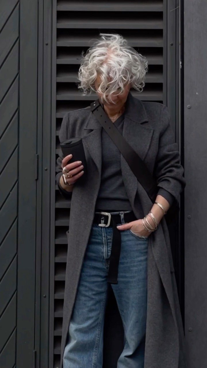 A person in a long grey coat and wide-leg blue jeans standing against a dark slatted wall.