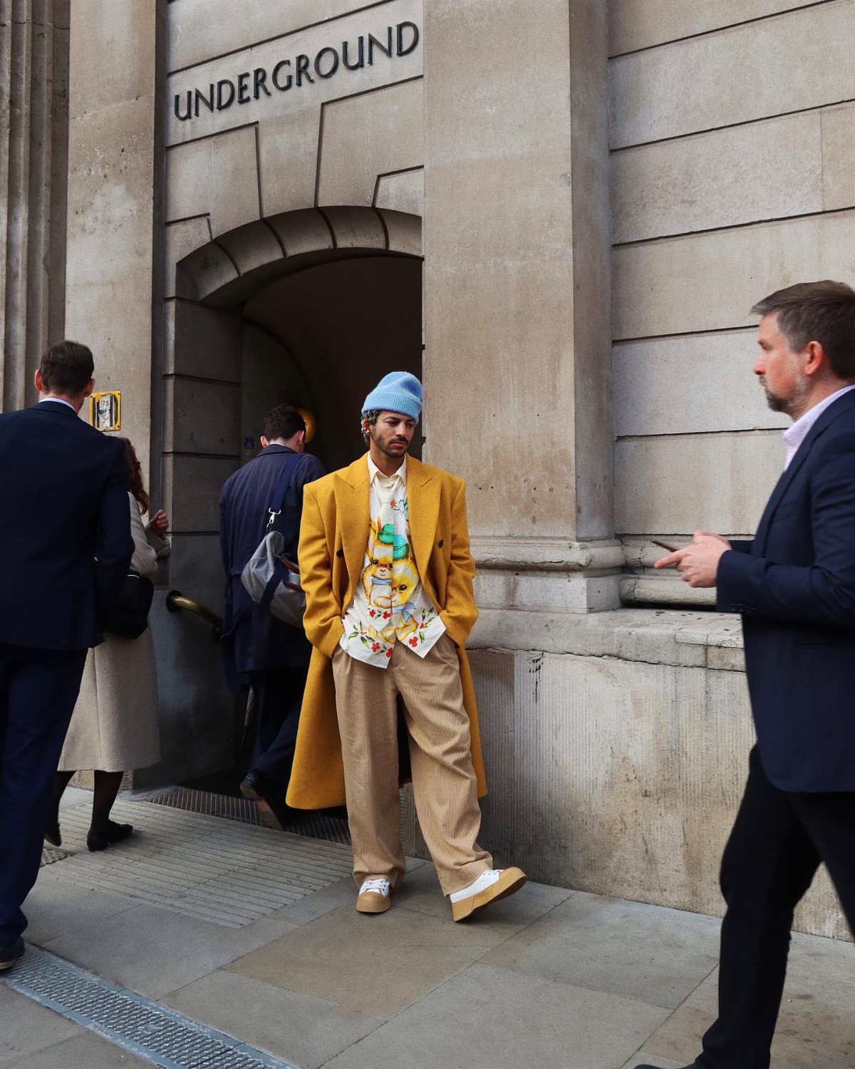Man in a bright mustard yellow oversized coat and tan trousers walking outside.