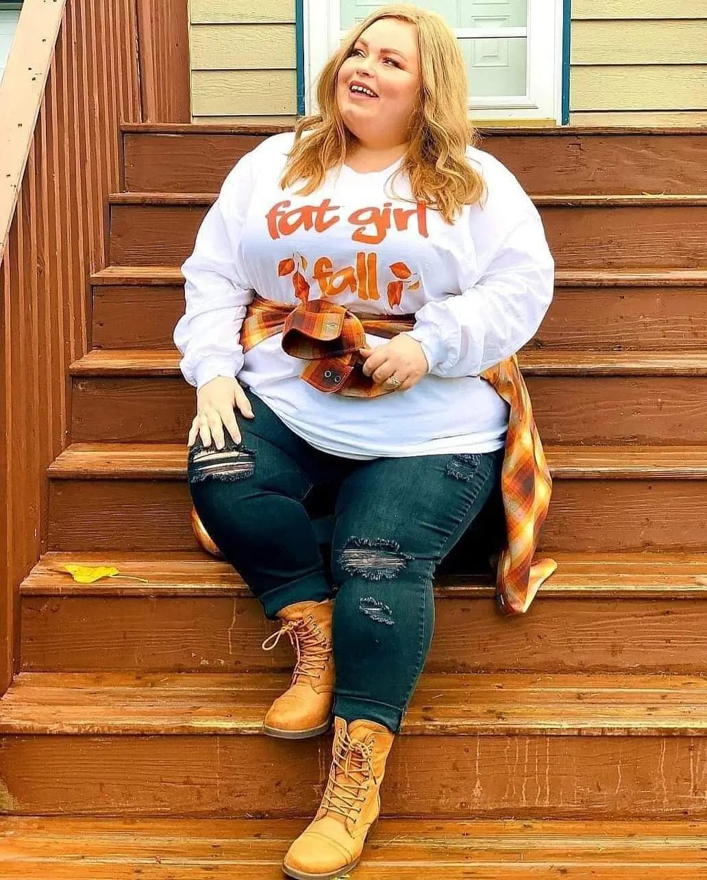 Plus size woman sitting on wooden stairs wearing a white sweatshirt and a flannel tied around her waist.