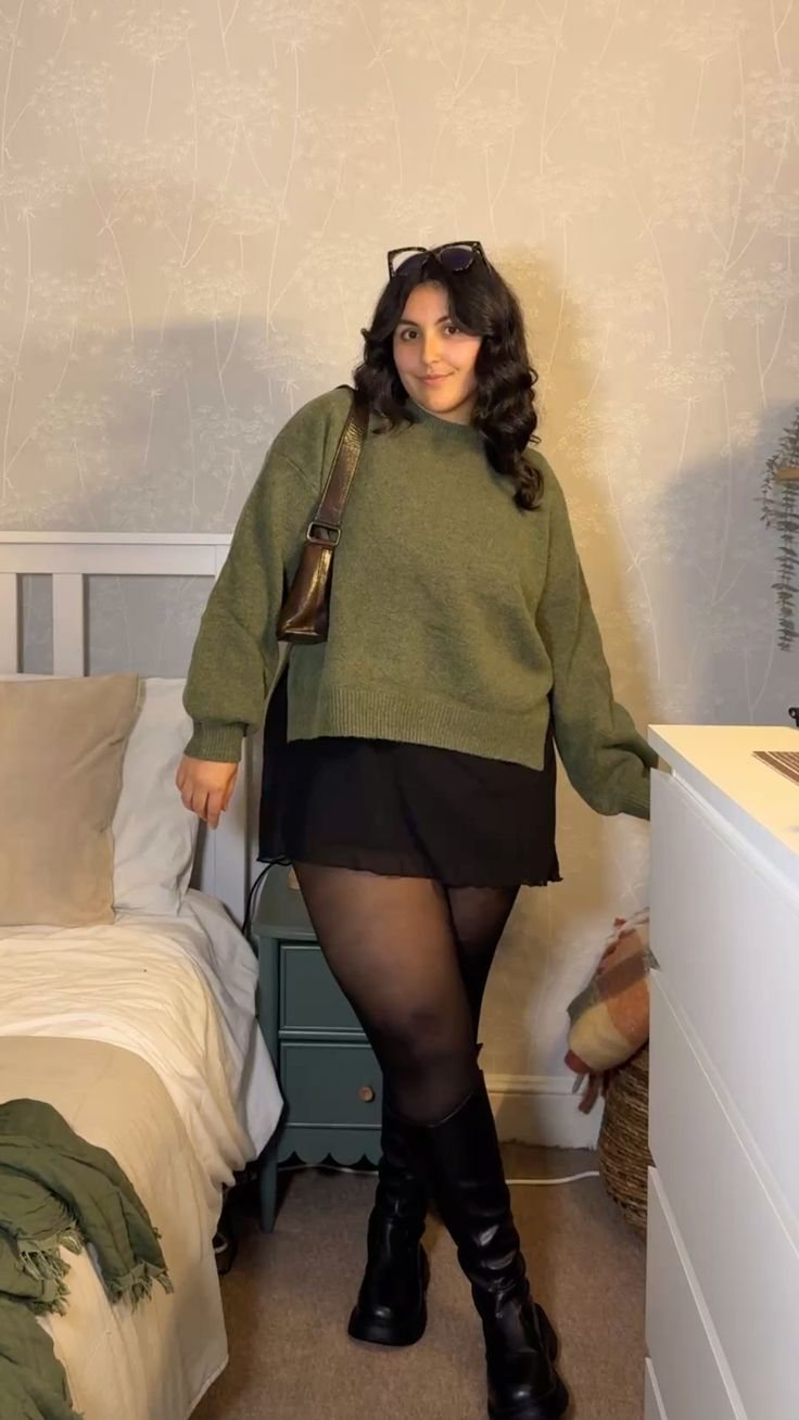 A woman wearing an olive green sweater, black skirt, and black knee-high boots standing in a bedroom.