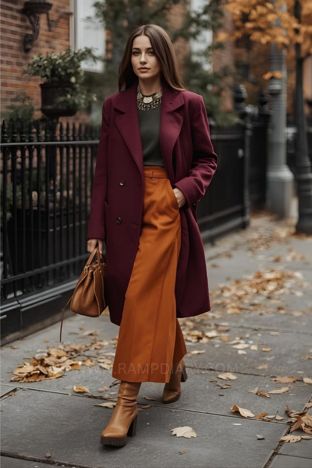 A woman walking outdoors wearing a burgundy coat, orange skirt, and brown boots on a leaf-strewn sidewalk.