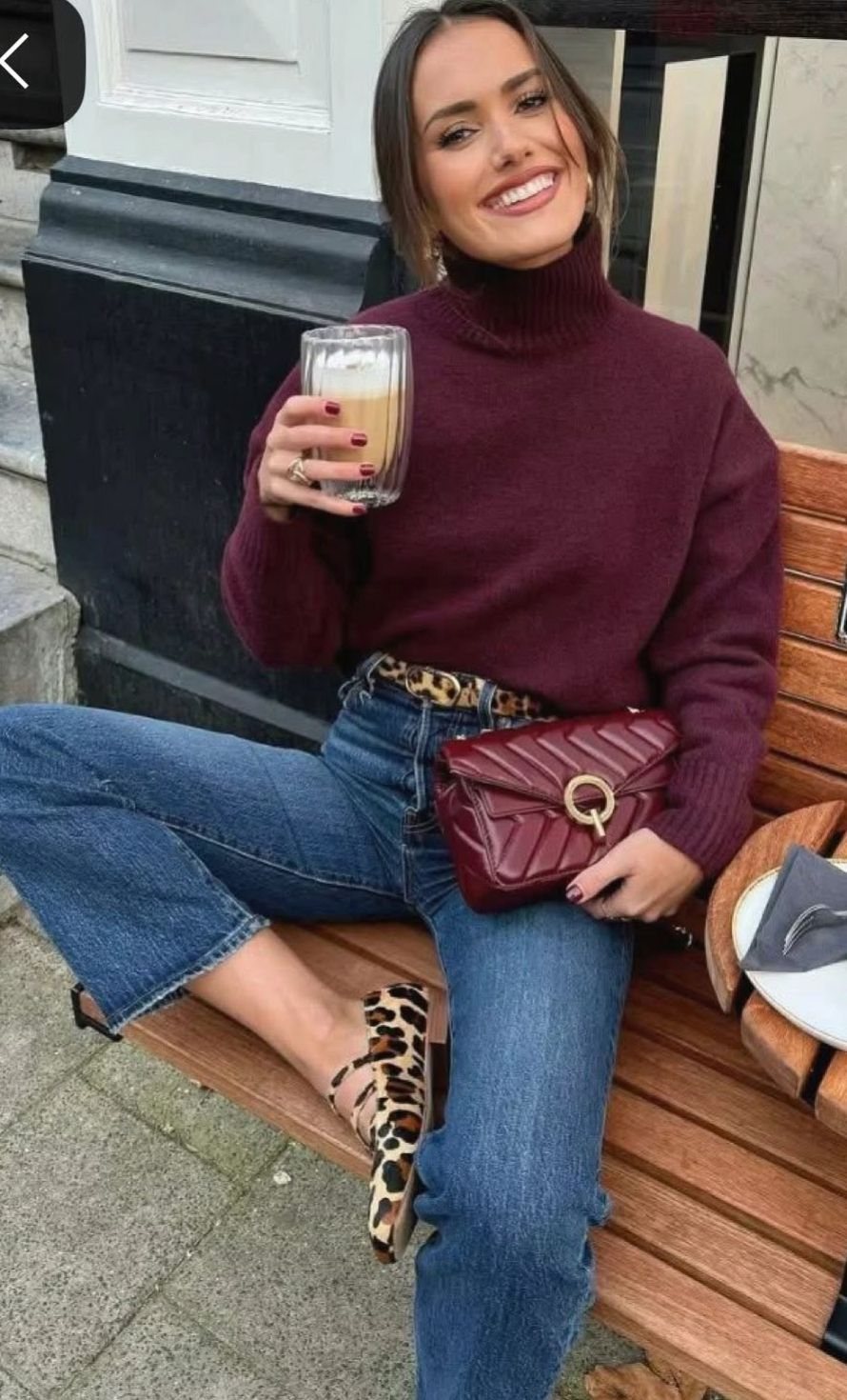Smiling woman in a burgundy turtleneck sweater and blue jeans sitting on a wooden bench.