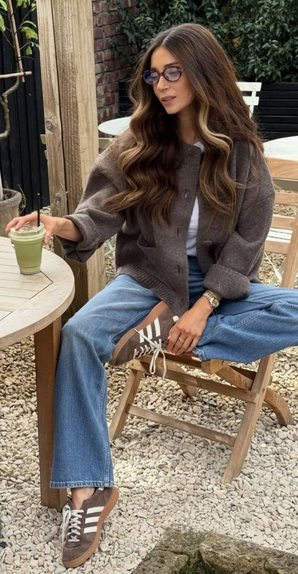 Woman in a brown cardigan and blue jeans sitting at an outdoor cafe table.