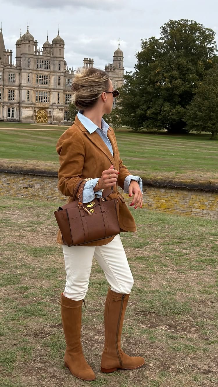 Woman wearing a tan suede blazer, white pants, and tall brown boots standing in a field.
