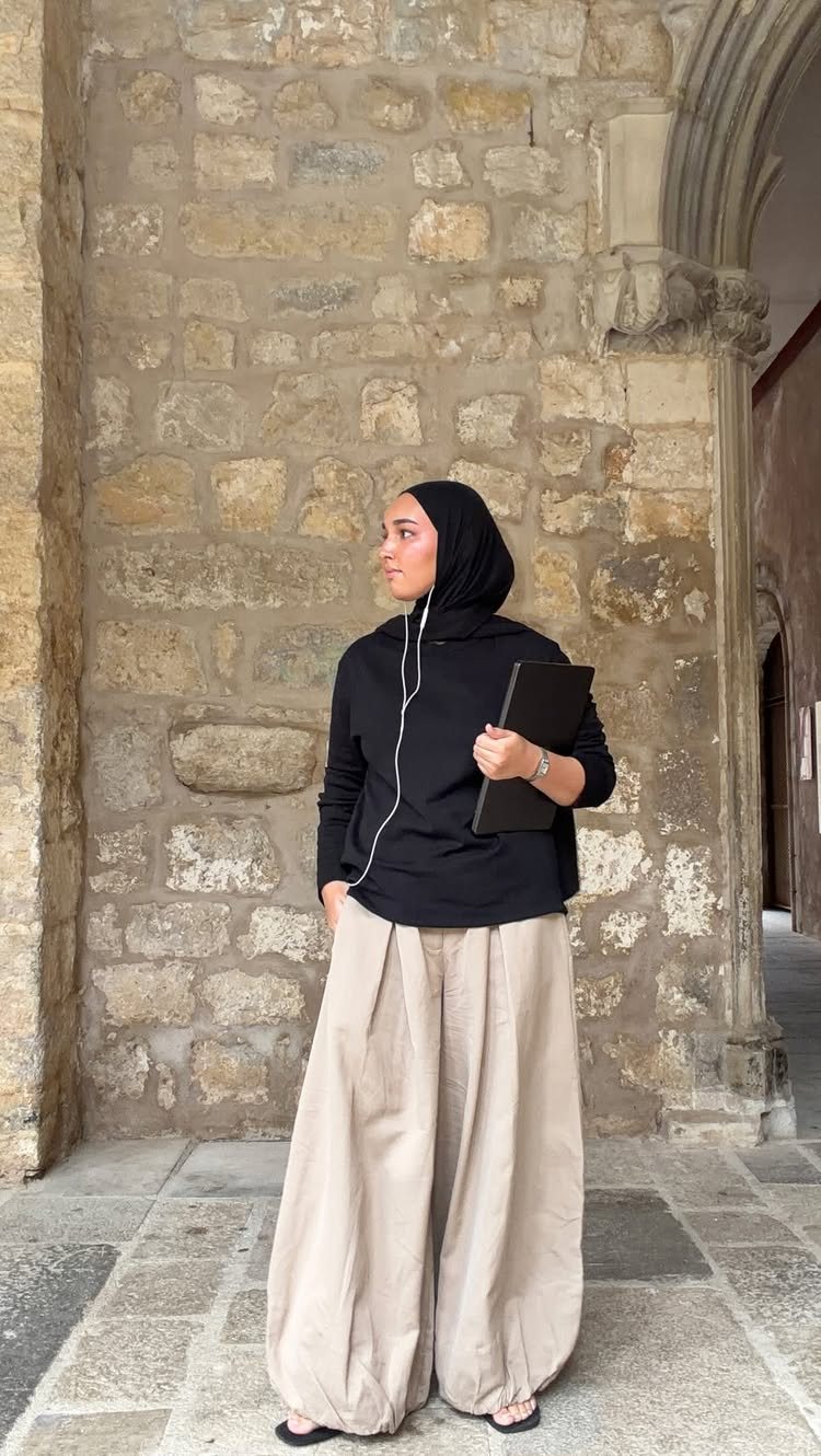 Woman in black hijab and black top wearing oversized beige balloon pants against a stone wall.