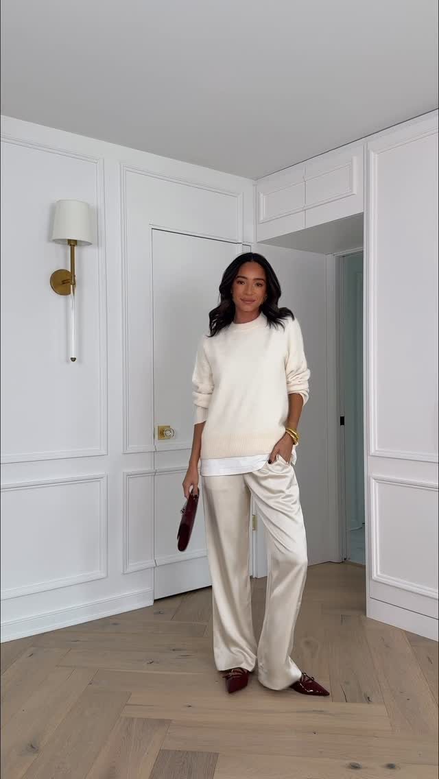 A woman posing in a white sweater and wide-leg white trousers in a bright room.