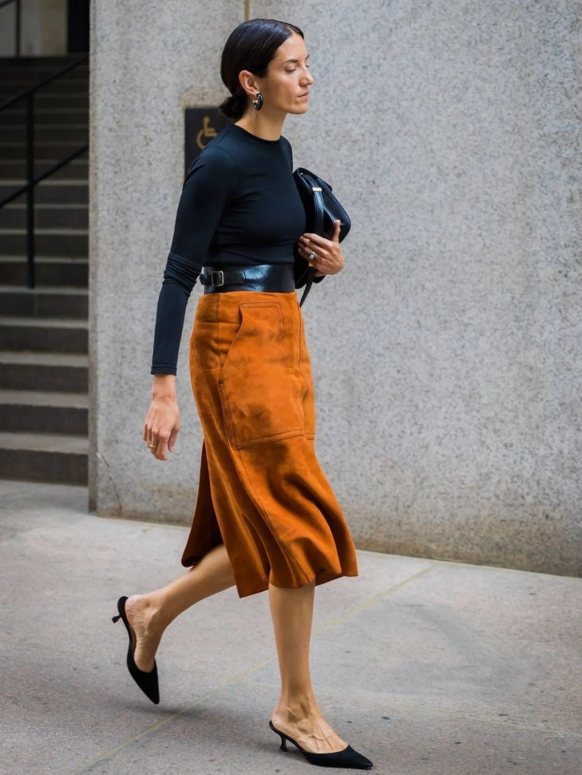 A woman walking in a black top and a brown suede midi skirt with black heels.