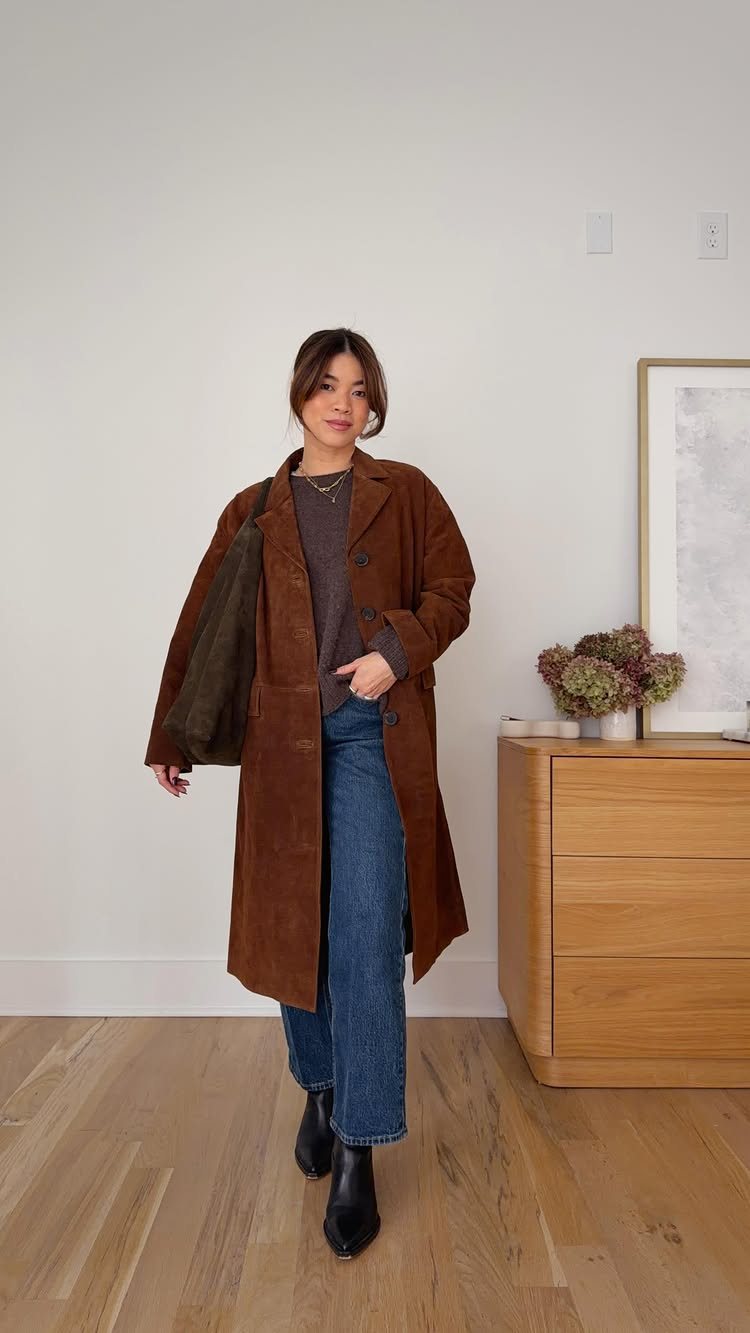 Woman wearing a long brown suede coat, blue jeans, and carrying a dark green suede handbag.