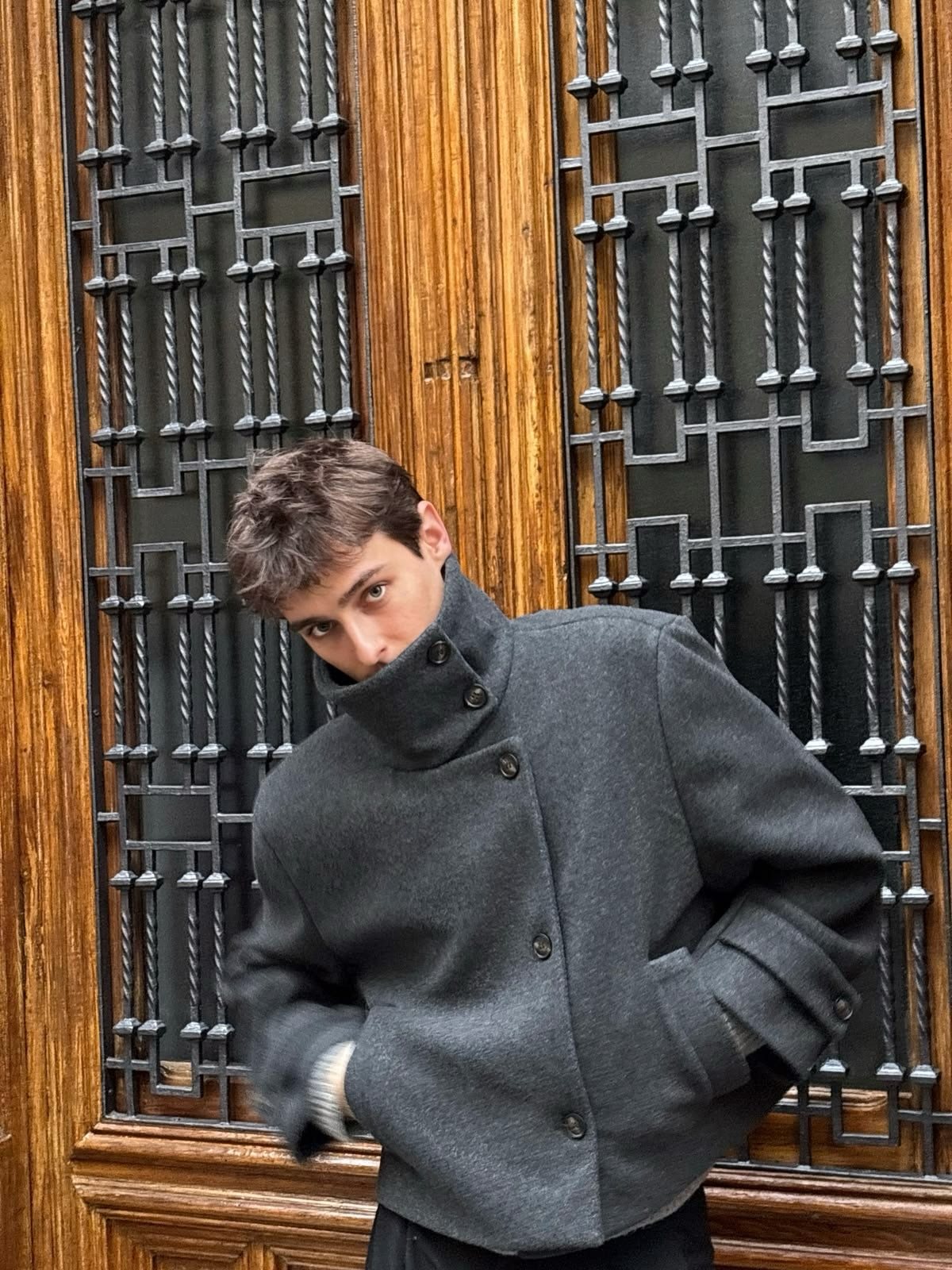 A young man posing in a dark grey high-neck jacket against a wooden door.