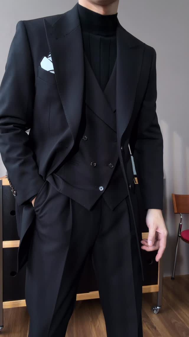 A person wearing a black formal suit with a matching black turtleneck and white pocket square.