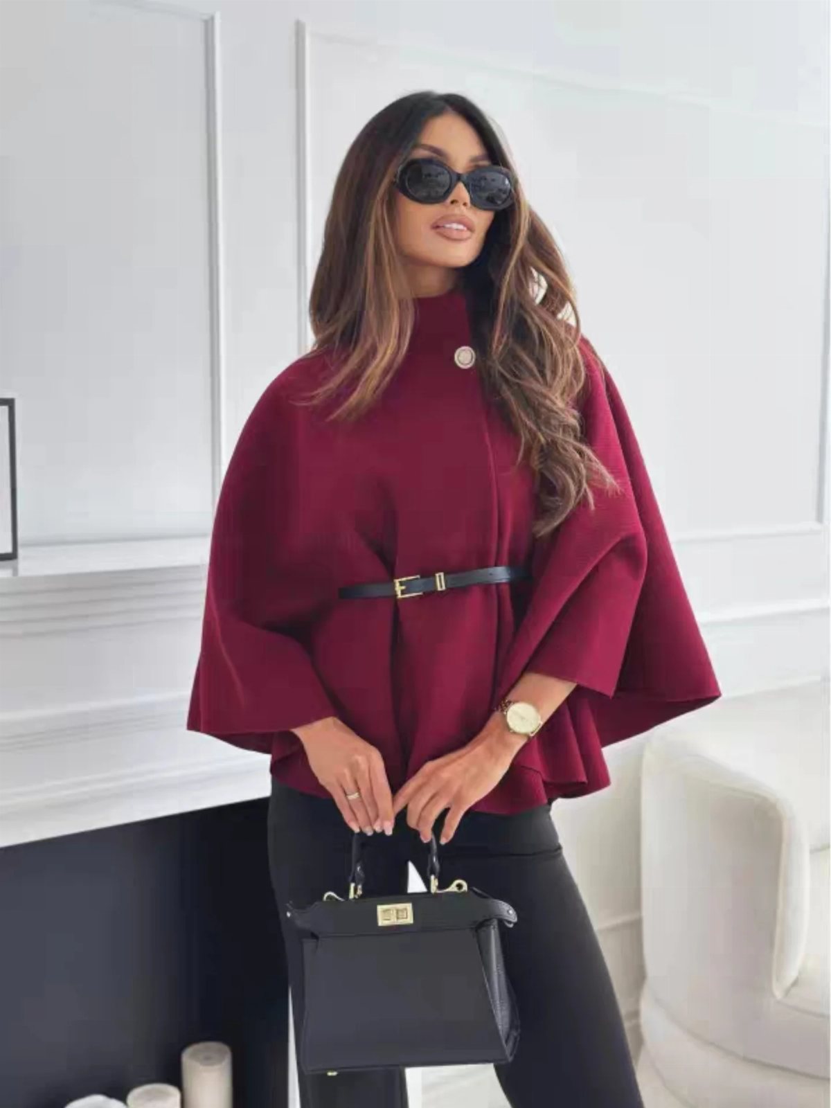 A woman posing in a burgundy cape coat cinched with a black belt and holding a handbag.