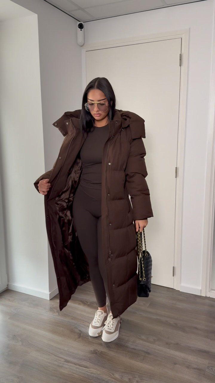 A woman posing in a long brown quilted puffer coat and matching brown outfit.