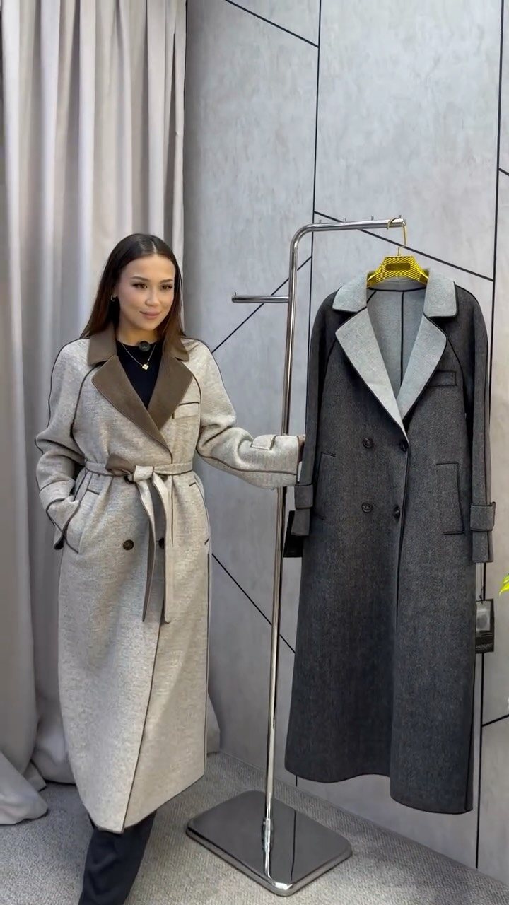 A woman standing beside a rack holding two long, structured wool coats in grey and charcoal.