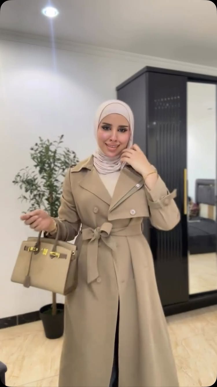A woman in a beige hijab and a long tan trench coat holding a handbag.