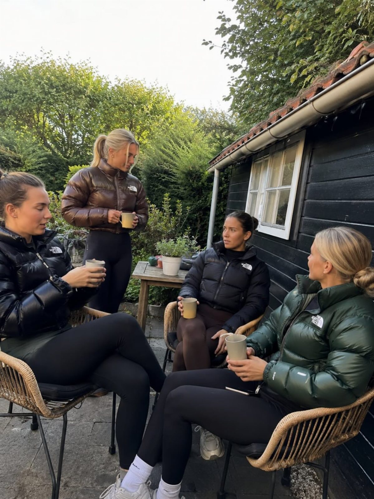 Four women sitting outdoors in puffer jackets while holding coffee cups in a garden.