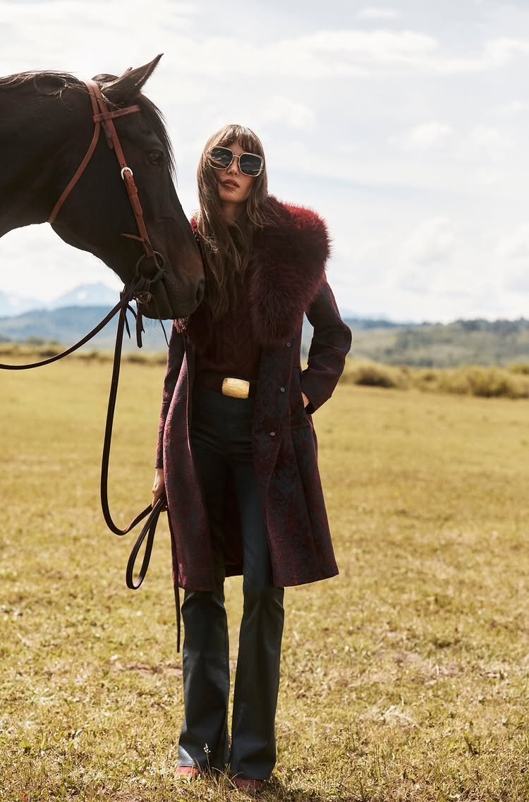 A woman in a burgundy fur-collared coat stands next to a horse in a field.