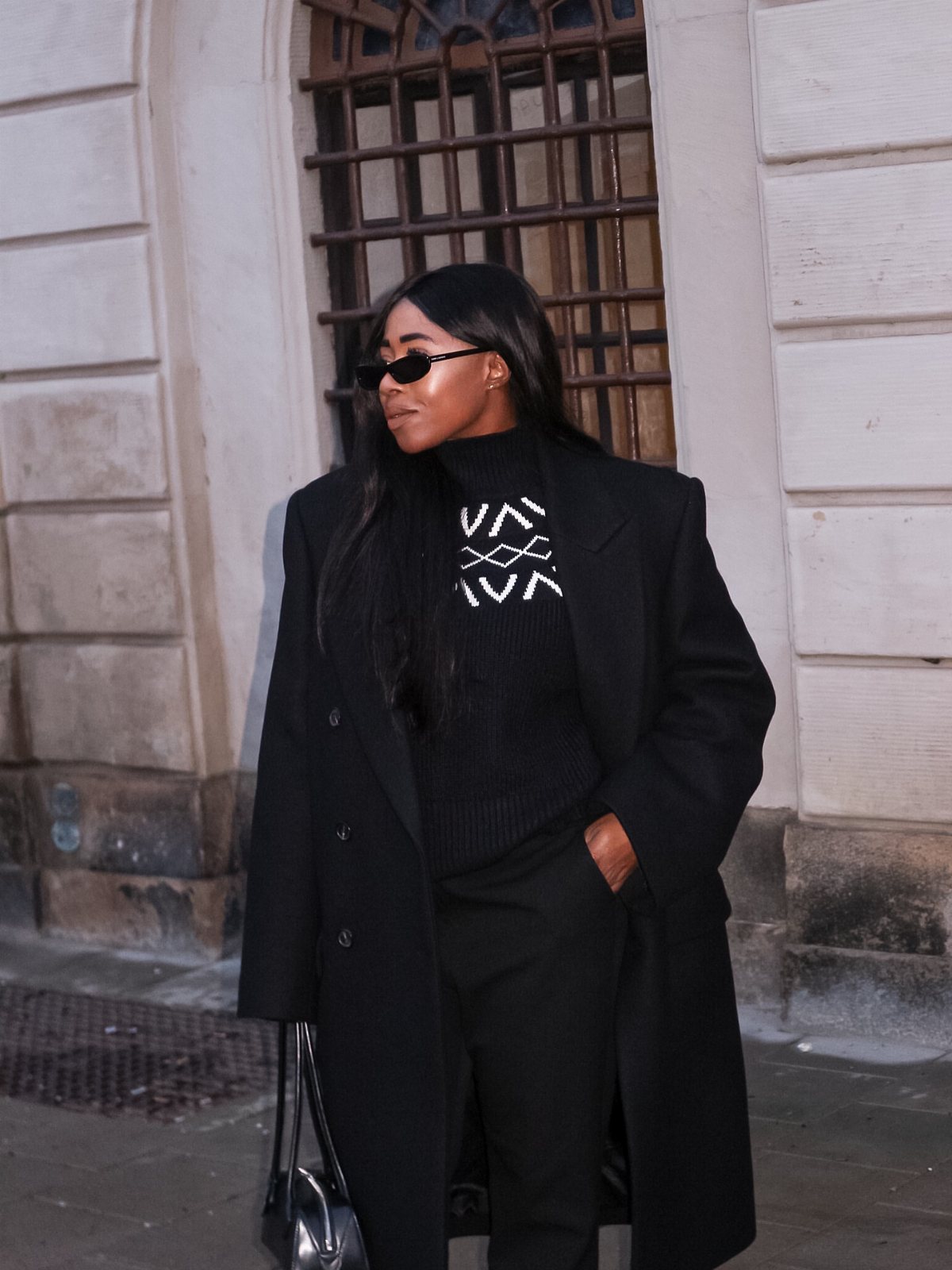 Woman in a black patterned turtleneck sweater and long coat standing outdoors.