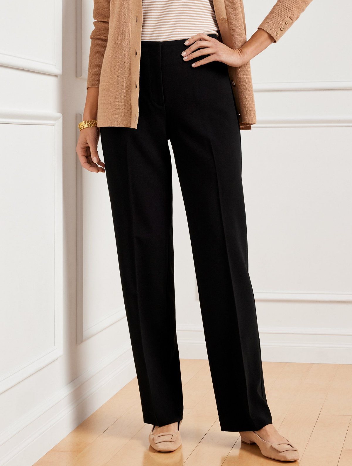 Woman wearing black wide-leg trousers, a tan cardigan, and beige flats against a white wall.