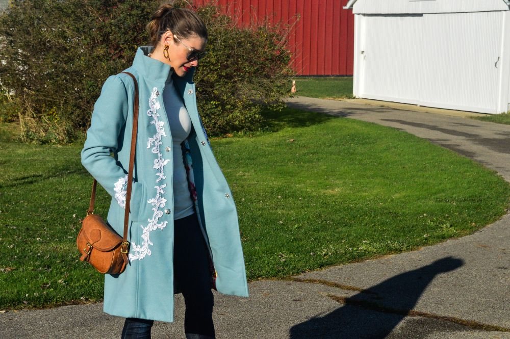 Woman walking in a long light blue coat with white floral embroidery and a high collar.