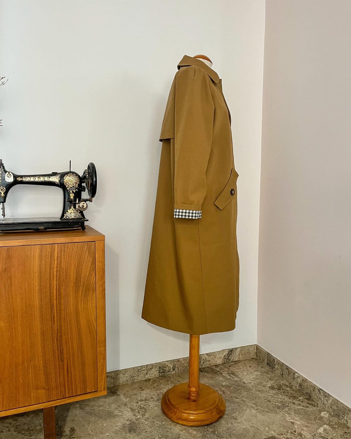 A long mustard-yellow coat with checkered cuffs displayed on a wooden mannequin stand near a vintage sewing machine.