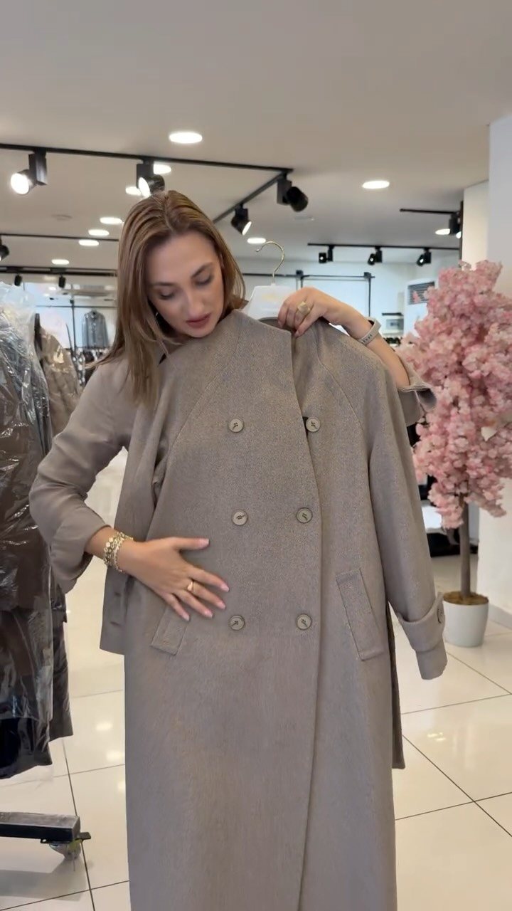 A woman holding up a long, taupe double-breasted wool coat in a retail store.