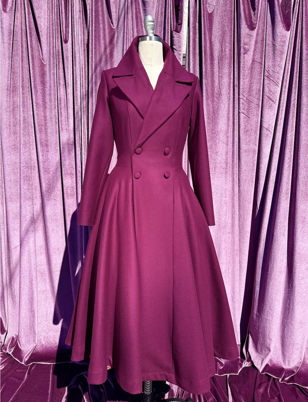 Magenta flared double-breasted coat with wide lapels displayed on a mannequin against purple curtains.