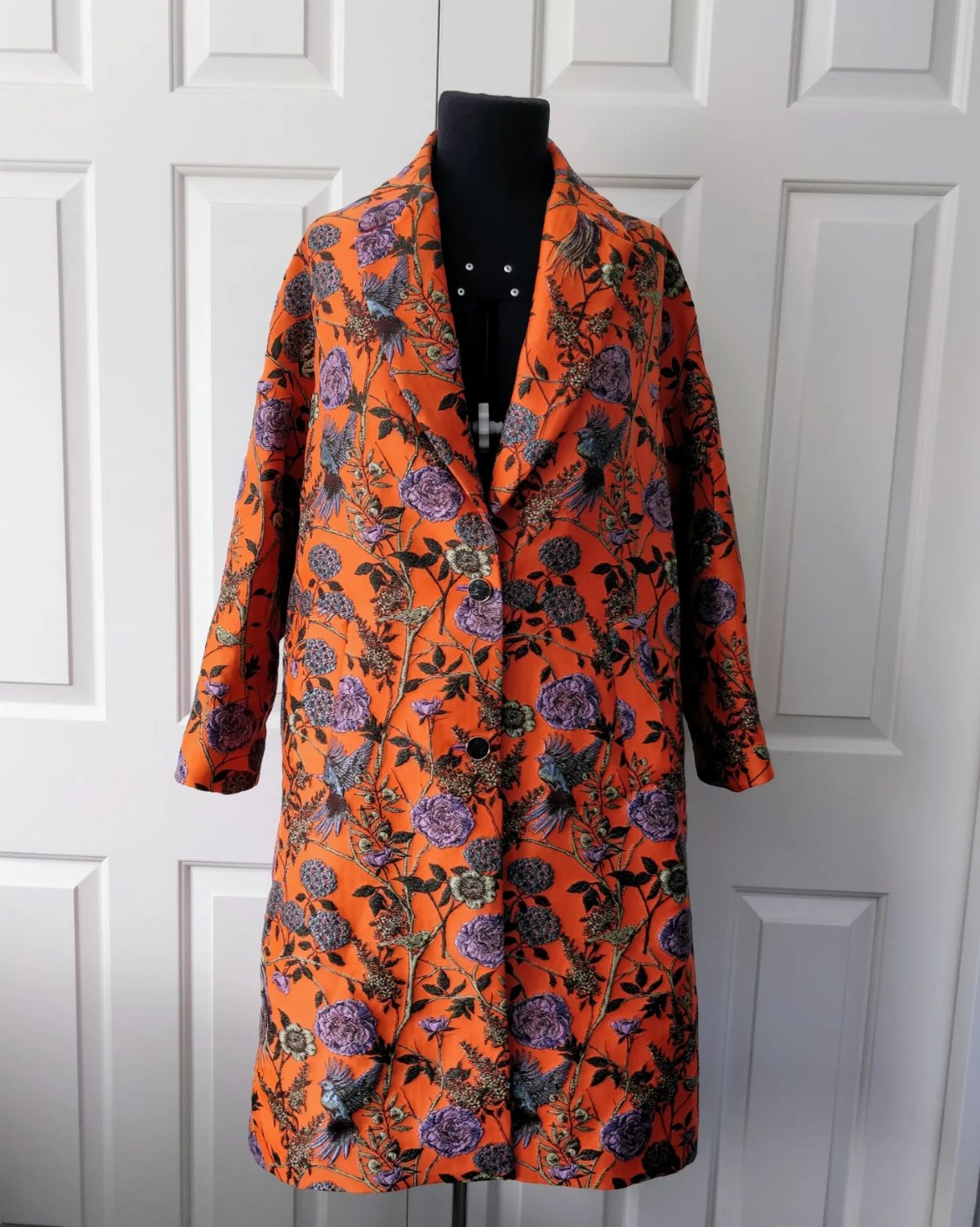 Orange floral patterned coat displayed on a black mannequin against a white door.