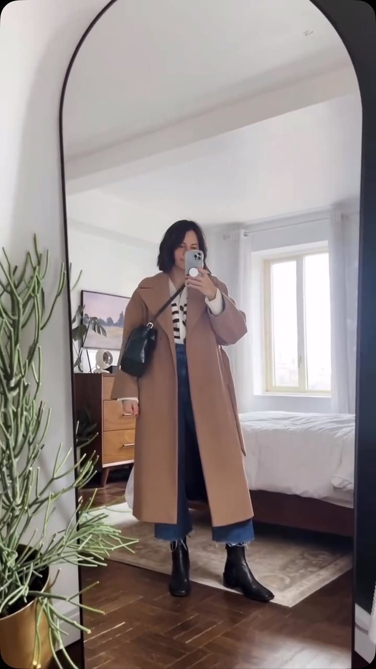 Woman posing in a mirror wearing a long camel wool coat, striped shirt, and blue jeans.