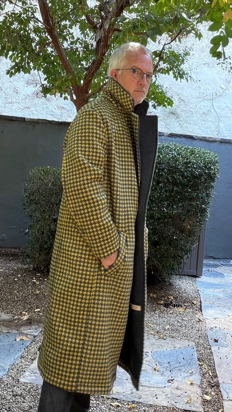 A man in a yellow and black houndstooth wool coat standing outdoors near greenery.