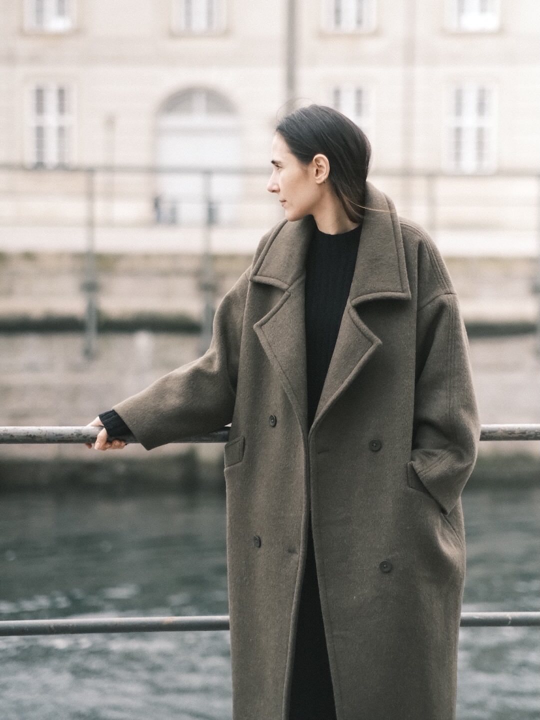 Woman wearing an oversized olive green wool coat standing by a metal railing near water.