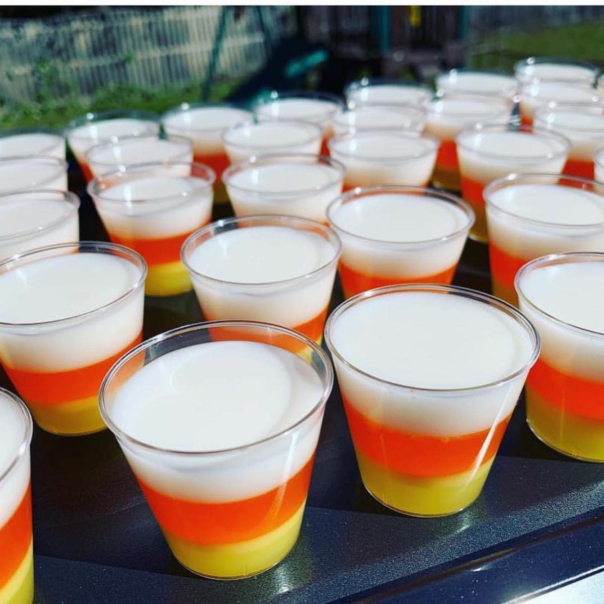 Candy Corn Jello Shots: 4 Layered Recipes for Halloween 1 Layered candy corn jello shots in clear plastic cups arranged on a dark surface.