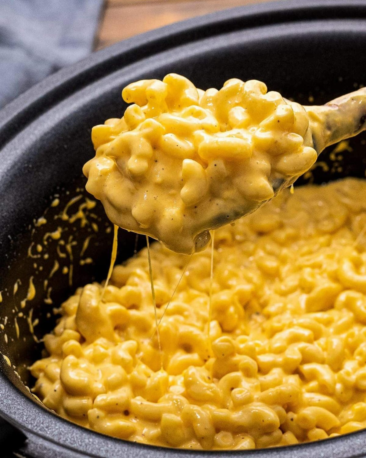 A metal spoon lifting creamy macaroni and cheese from a black slow cooker pot.