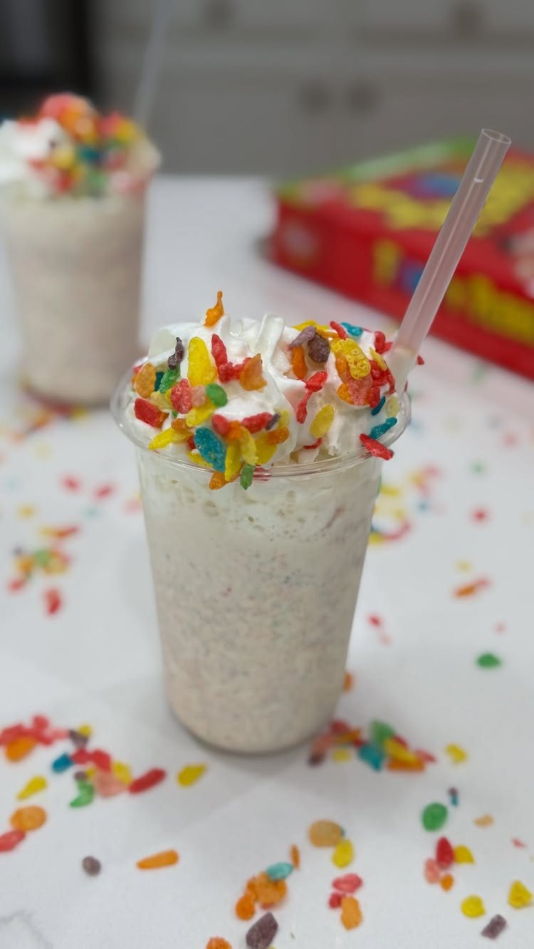 Two milkshakes topped with whipped cream and colorful fruity cereal on a white surface.
