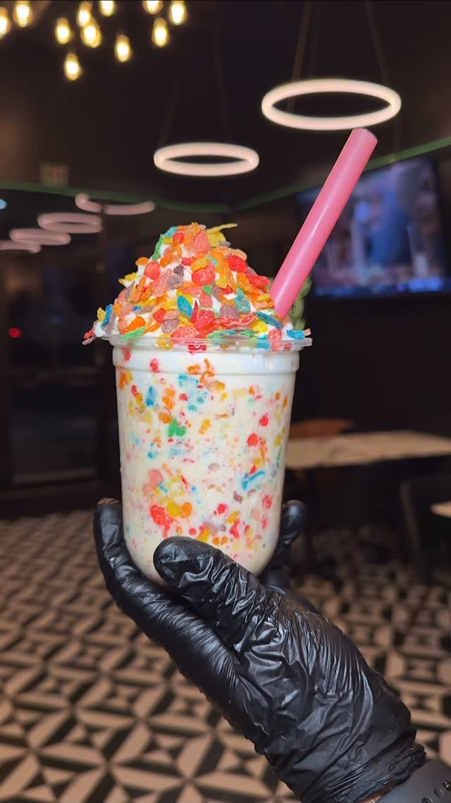 A Fruity Pebbles milkshake in a clear cup with a pink straw held by a gloved hand.