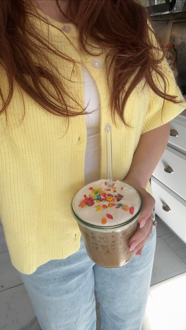 A person holding a glass of iced milkshake topped with colorful fruity cereal pieces.