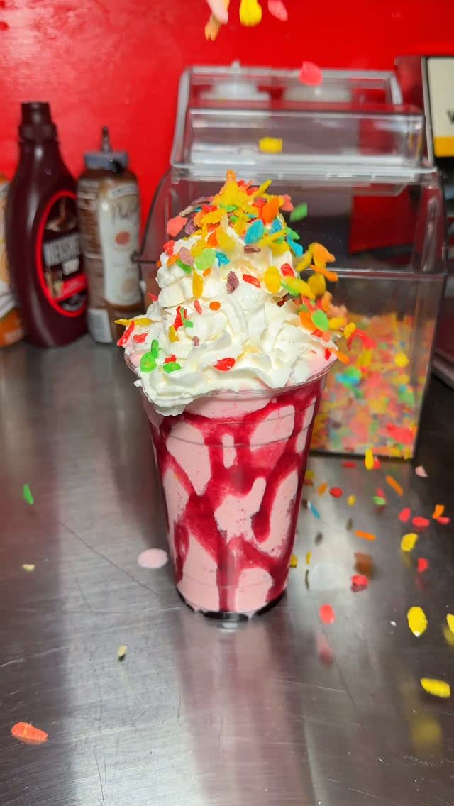 A pink milkshake topped with whipped cream and colorful fruity cereal pieces on a metal counter.