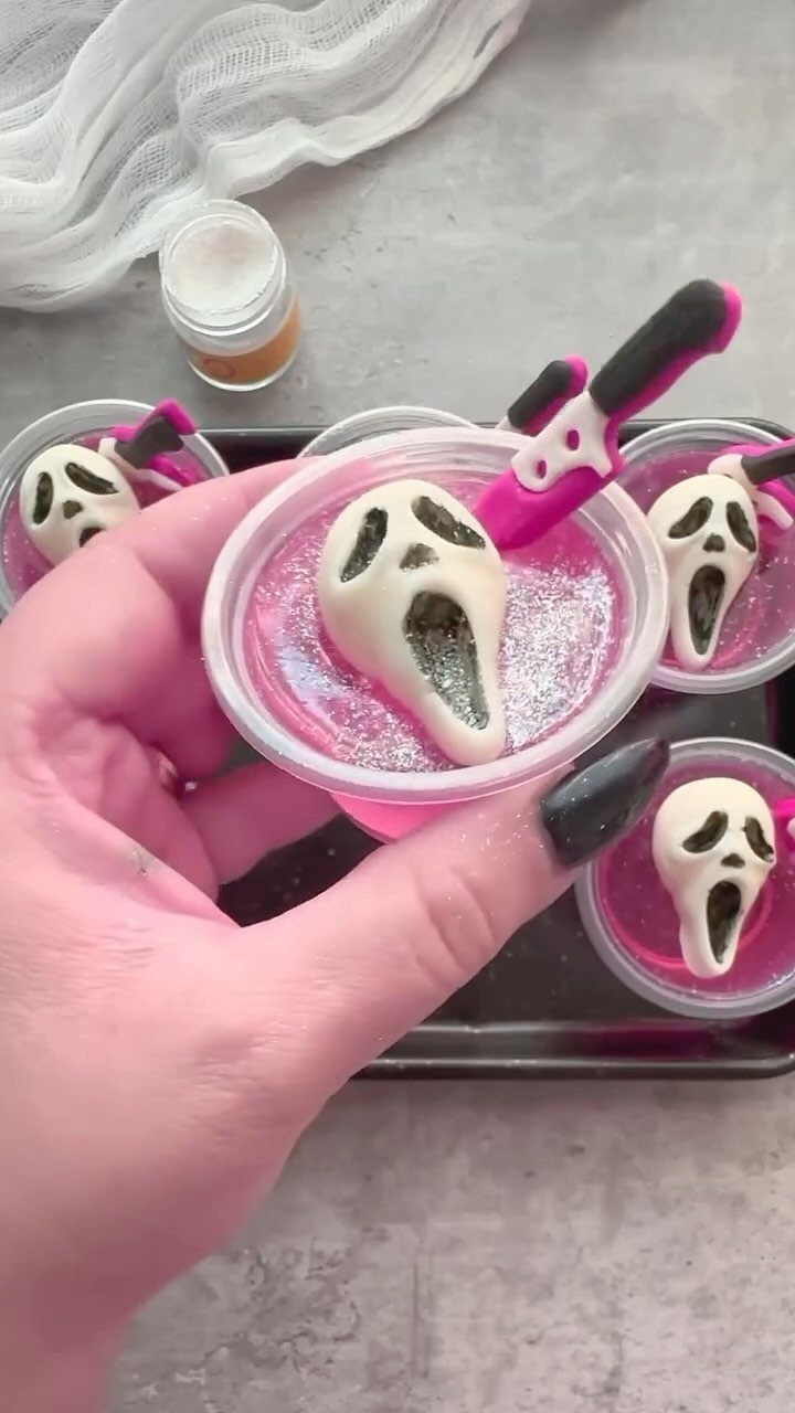 A hand holding a pink jello shot cup with a white Scream mask shaped gummy candy.