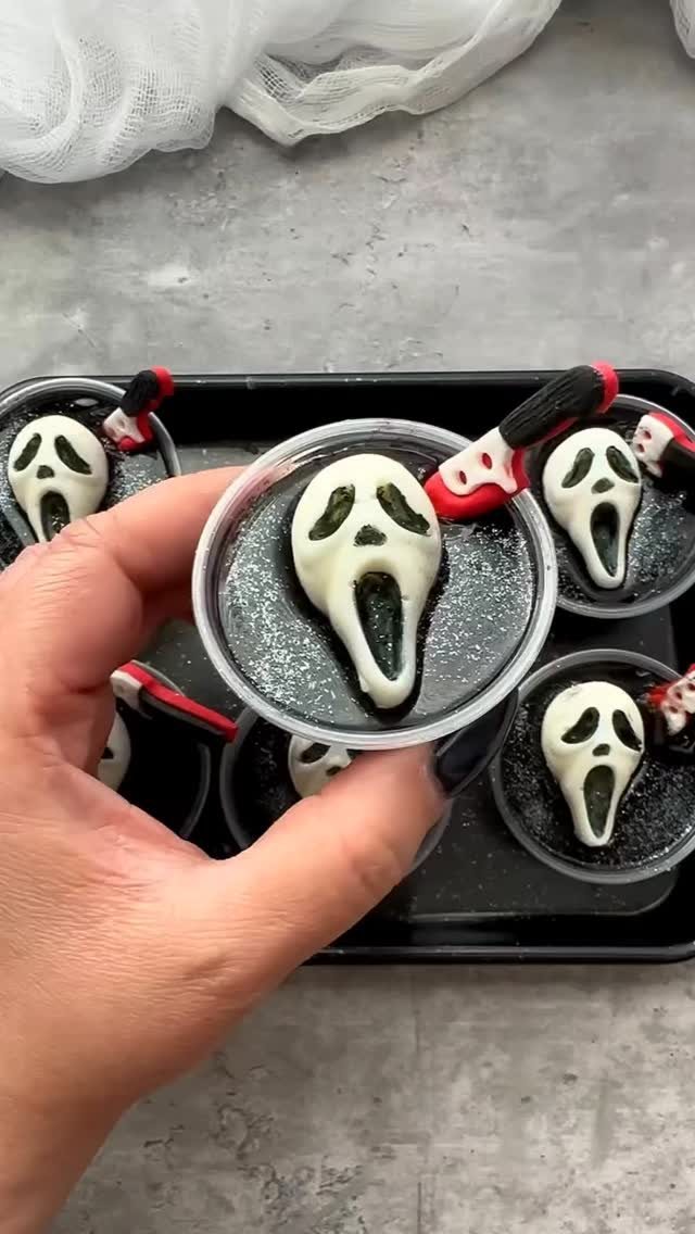 A hand holding a dark jello shot topped with a white ghost mask decoration.
