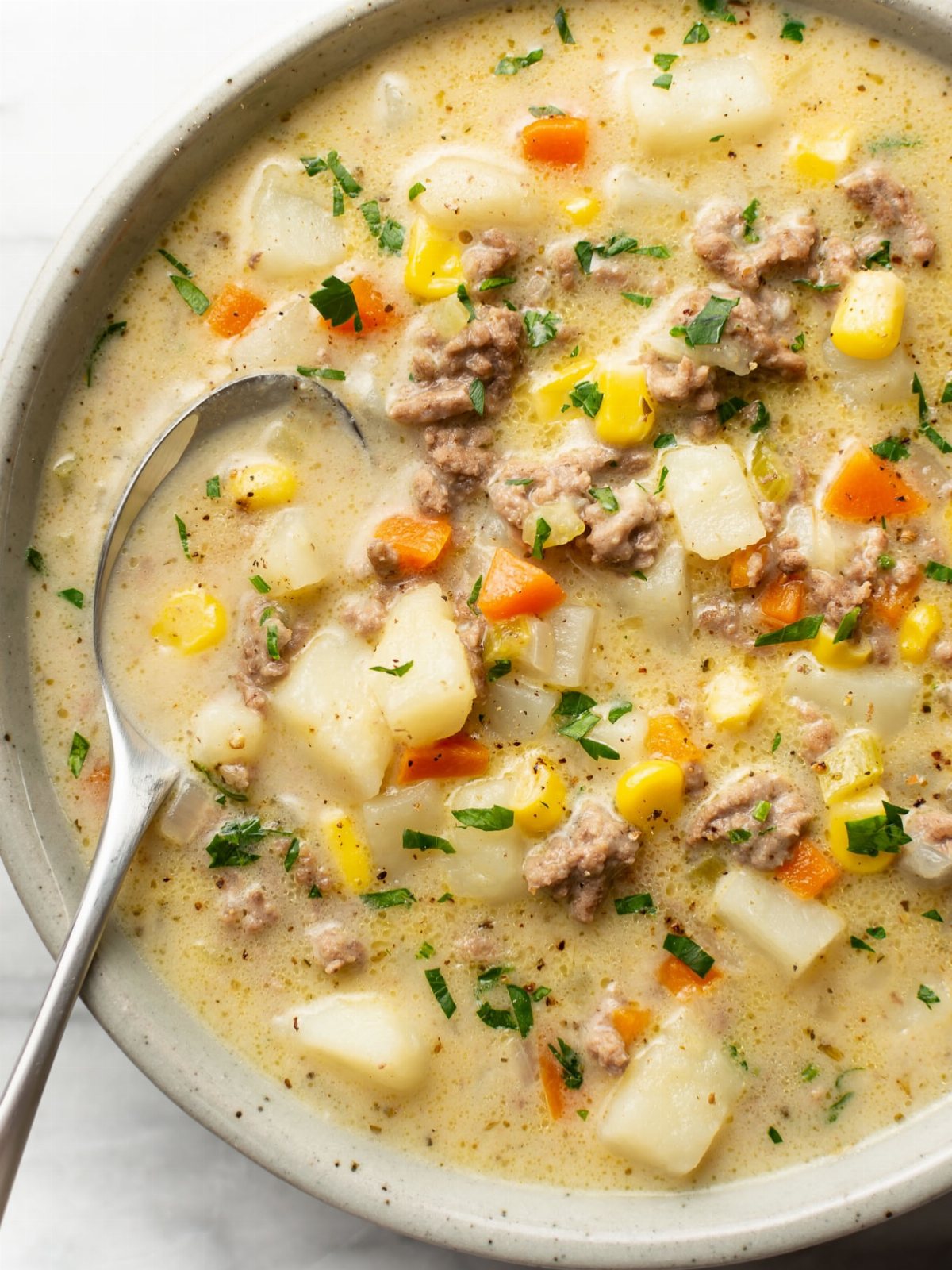 14 Ground Beef Recipes Using Few Ingredients for Low-Stress Weeknights 10 Creamy hamburger soup with ground beef, potatoes, corn, and carrots in a white bowl with a spoon.