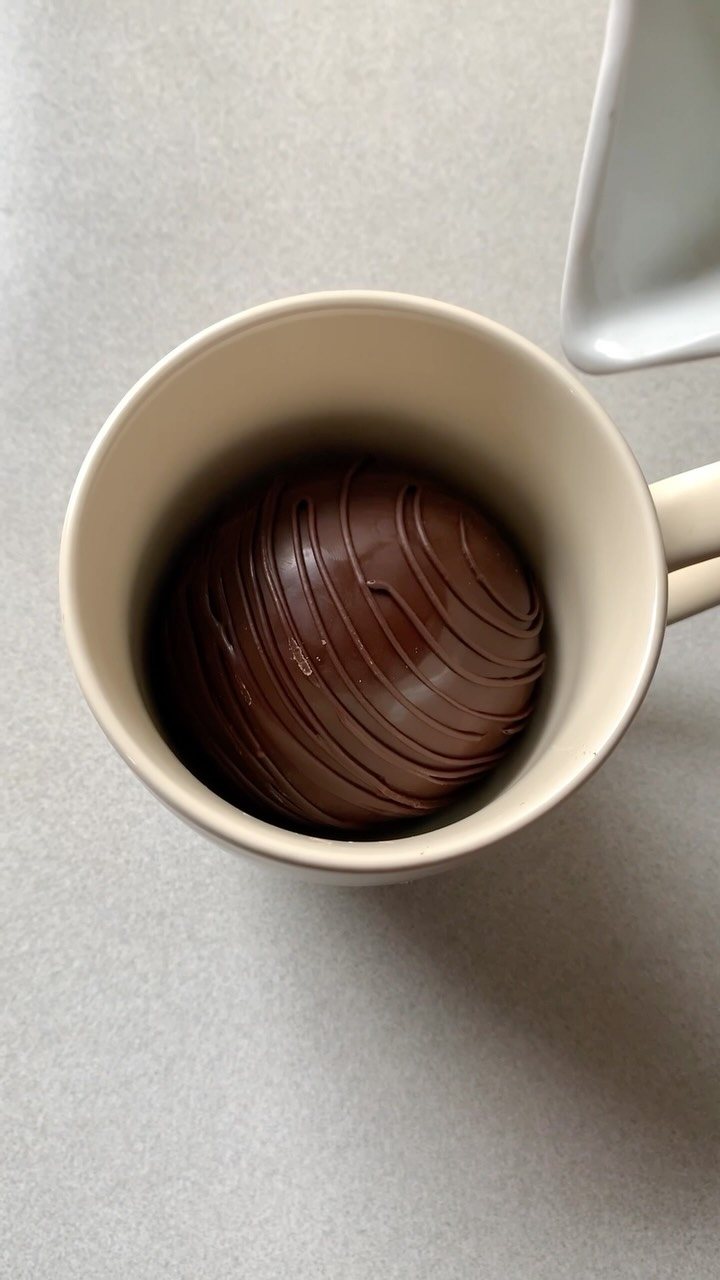12 Hot Chocolate Bombs for Every Holiday and Skill Level 1 A chocolate bomb with drizzled lines resting inside a cream ceramic mug.