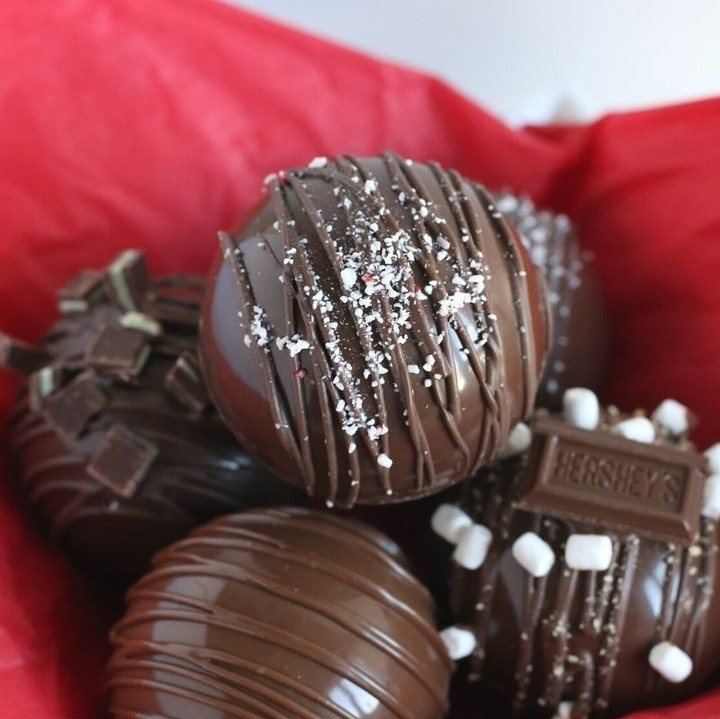 12 Hot Chocolate Bombs for Every Holiday and Skill Level 3 Chocolate hot chocolate bombs with peppermint sprinkles and chocolate drizzle in red tissue paper.