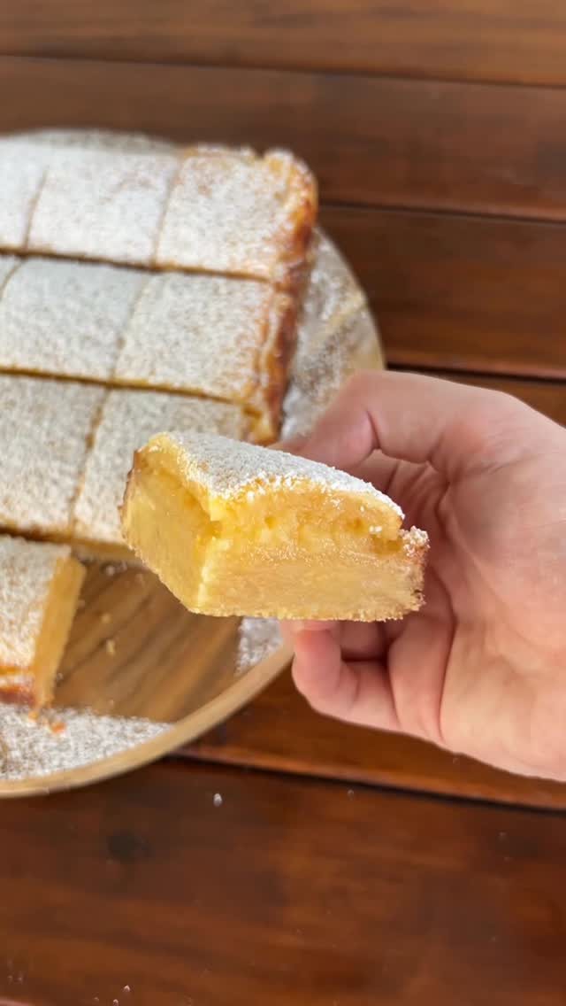 A hand holding a single piece of powdered lemon bar over a plate of sliced bars.