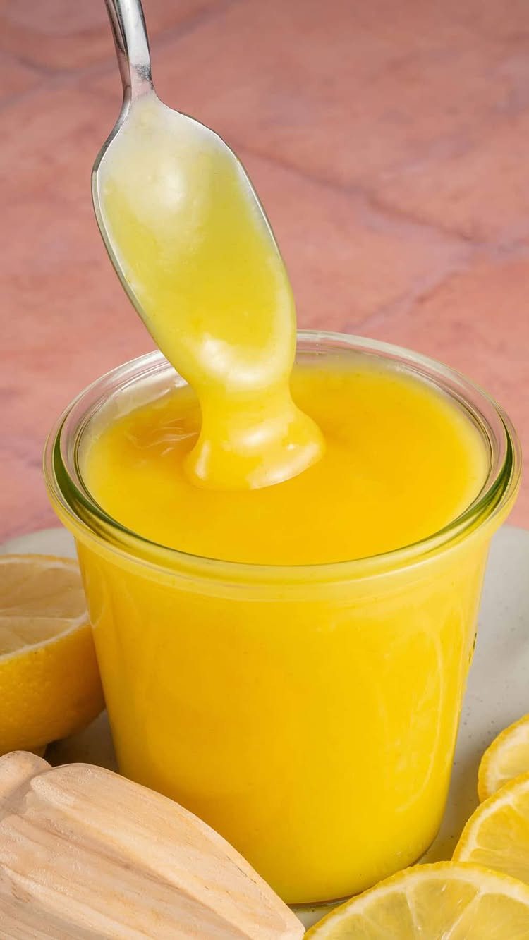 A silver spoon lifting thick, bright yellow lemon curd from a clear glass jar.