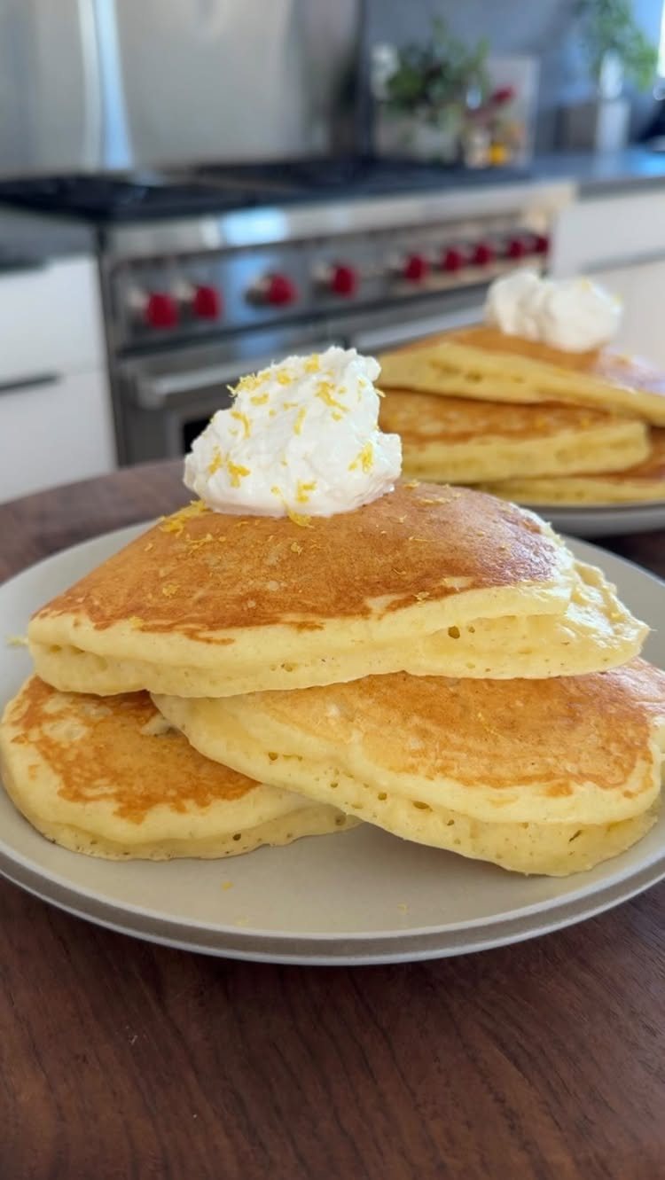 Stack of fluffy golden pancakes topped with white cream and lemon zest on a white plate.