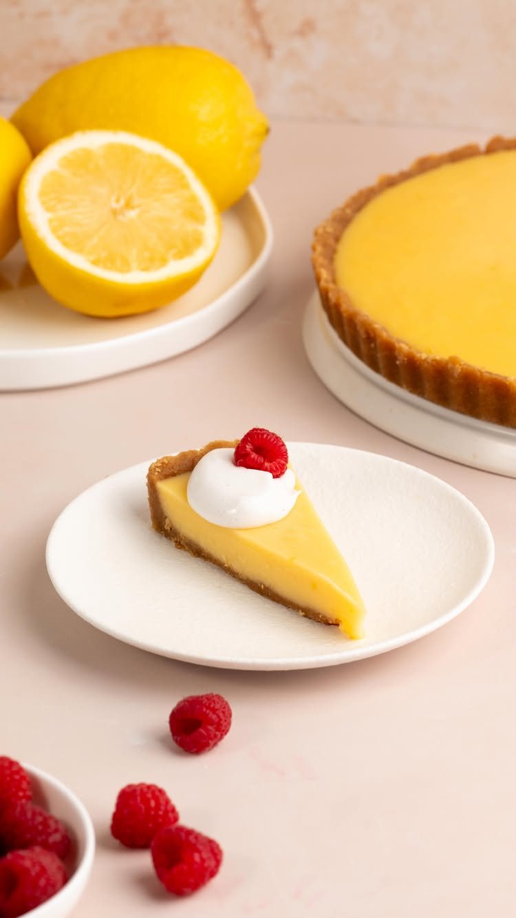 A slice of lemon tart with whipped cream and a raspberry on a white plate.