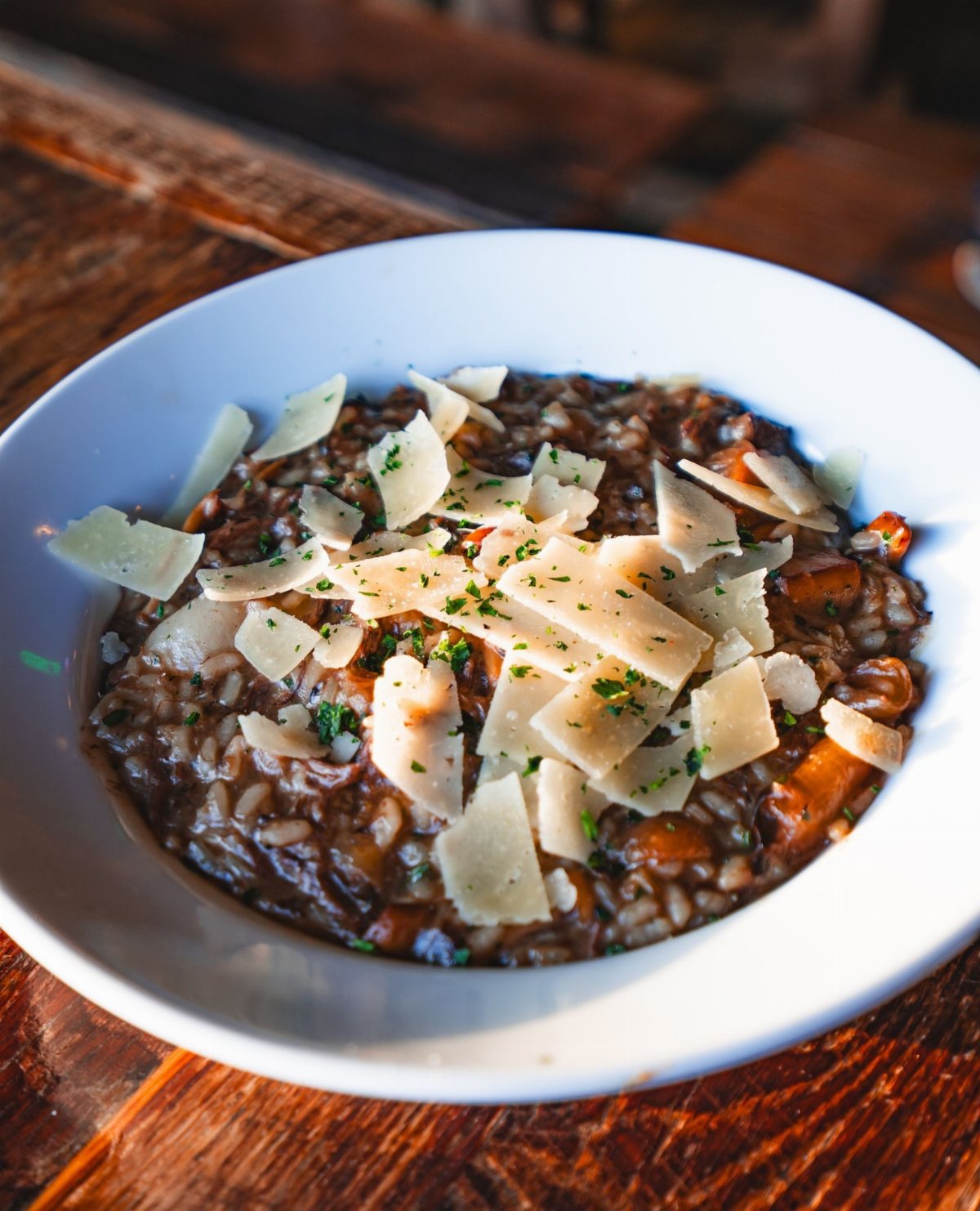 Creamy mushroom risotto topped with large parmesan cheese shavings and fresh green herbs in a white bowl.